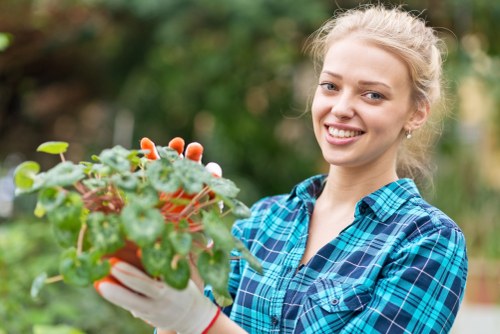A young woman with blonde hair tied back, wearing a blue and green plaid shirt and gardening gloves, is smiling while holding a vibrant potted plant with green and reddish leaves in a garden. The outdoor space features a well-maintained lawn with dense, lush grass, and a neatly landscaped border with shrubs and small trees in the background. The garden appears to be part of a residential property in Forest Gate, with natural daylight illuminating the scene, indicating a clear or partly cloudy day. This setting illustrates typical elements of a landscaped garden suitable for professional gardening and landscaping services offered by Landscapers Forest Gate, including flower bed creation and outdoor plant maintenance.