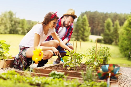 In a well-maintained outdoor garden space, a man and a young girl are tending to a raised vegetable bed, with the girl wearing a pink headband and the man wearing a straw hat and a plaid shirt. The garden features a variety of small plants and seedlings, some supported by bamboo stakes, with the soil appearing moist and dark. Adjacent to the vegetable bed, there is a small garden decoration in the shape of a flower. The background showcases a lush, green landscape with tall trees and open grassy areas under a bright, sunny sky, indicating clear weather conditions. The overall setting illustrates a peaceful, cultivated outdoor environment typical of a residential garden in Forest Gate, with a focus on gardening and outdoor maintenance, aligned with professional landscaping services offered in the area.