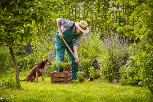 A person wearing a wide-brimmed straw hat, a blue checked shirt, and green overalls is tending to a garden on a lush, green lawn. They are using a small hand tool to work on the soil near a wooden planter box filled with potted plants. To the left, a brown dog is sitting attentively, observing the gardener. The garden is surrounded by a variety of tall plants, shrubs, and flowering perennials, with dense green foliage providing a natural backdrop. The lawn area features well-maintained, vibrant grass with a gentle slope, bordered by trees and bushes. The scene is set outdoors during daylight, with soft natural light illuminating the garden's diverse textures and colours, creating an inviting, peaceful environment suitable for gardening and outdoor maintenance services often offered by companies like Landscapers Forest Gate in the E6 postcode area of London.