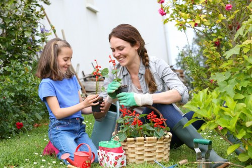 A woman and young girl are outdoors in a lush garden with vibrant green foliage, shrubs, and flowering plants. They are seated on the grass, engaging in gardening activities with smiles. The woman is wearing green gardening gloves and is holding a small potted plant with red flowers, while the girl, dressed in a blue T-shirt and denim overalls, is also holding the same type of pot. Nearby, there is a woven basket filled with gardening tools and materials, including a red watering can and a container with a festive design. Behind them, various garden borders, including bushes and a hedge, frame a well-maintained outdoor space, suggesting a peaceful and organized landscaped garden typical of residential properties in Forest Gate, London. The scene depicts a calm, sunny day suitable for gardening, with soft natural light illuminating the scene, emphasizing the natural tones of green and red, and highlighting the connection to local gardening and landscaping services like those offered by Landscapers Forest Gate in the E7 postal district.