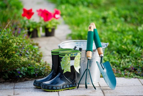 The image shows a close-up of a garden pathway with gardening tools and accessories laid out in the foreground. There are a pair of black and green rubber gardening boots positioned on the stone paving, with a cloth draped over one of the boots. Next to the boots, there are a pair of gardening gloves hanging from a bucket, which is placed on the paving and appears to be filled with soil or compost. A small hand trowel with a blue handle rests against the bucket, and a garden fork with green handles is leaning beside it. In the slightly blurred background, vibrant pink and red flowers are visible, along with lush green foliage and a well-maintained lawn or garden borders. The setting appears to be a tidy outdoor space, possibly in a front or back garden in Forest Gate, with natural lighting suggesting a clear, bright day. This scene captures essential gardening tools and outdoor plant-care elements, relevant to landscaping and garden maintenance services available locally from Landscapers Forest Gate, supporting garden upkeep and outdoor improvements in the area.