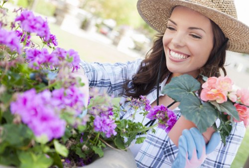 A woman wearing a straw sunhat and blue gardening gloves is outdoors in a landscaped garden area, tending to a variety of flowering plants arranged in pots and planter beds. The garden features a mix of vibrant purple and pink flowers with green foliage, situated on a paved patio or terrace. Behind her, a well-maintained lawn or grassy patch is visible, bordered by shrubs or trees that provide shade and natural backdrop. The weather appears bright and sunny, contributing to the lively atmosphere of gardening activity. The scene illustrates a neat, organized outdoor space suitable for relaxing or gardening services by Landscapers Forest Gate, with careful attention to plant arrangement and garden maintenance details typical of professional landscaping in the local area near Forest Gate, London.