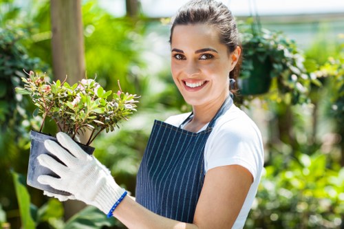 A young woman with dark hair tied back, wearing a white t-shirt, striped gardening apron, and white gardening gloves, is smiling while holding a potted plant with small green and purple-tinged leaves. She stands in a lush garden environment with vibrant green foliage, including trees, shrubs, and flowering plants, suggesting a healthy and well-maintained outdoor space. The background shows a blurred view of a garden shed or greenhouse, with natural sunlight illuminating the scene, highlighting the fresh growth and natural tones of the garden. This setting reflects a carefully landscaped area suitable for gardening or landscaping projects, such as creating flower beds or enhancing outdoor spaces, aligning with services offered by Landscapers Forest Gate. The overall scene captures the care and attention involved in garden maintenance and planting in the Forest Gate area, emphasizing natural beauty and professional outdoor landscaping practices.
