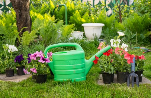 A garden scene showing a lush, green outdoor space with a variety of potted flowering plants, including pink, purple, and white blooms, neatly arranged around a well-maintained lawn. In the foreground, there is a bright green plastic watering can nestled among the plants, indicating ongoing gardening care. The background features mature trees with textured bark and leafy branches providing partial shade, along with dense foliage suggesting a private, landscaped garden area typical of a domestic back garden in Forest Gate. The scene appears to be set on a clear day with natural sunlight illuminating the vibrant colors and textures of the plants, soil, and lawn. The overall environment is tidy and inviting, reflecting professional gardening and lawn maintenance, with visible elements consistent with landscaping services offered by local experts like Landscapers Forest Gate. This outdoor space showcases a harmonious combination of natural plant growth and garden tools, suitable for enhancing a garden's aesthetic and health through professional gardening services.