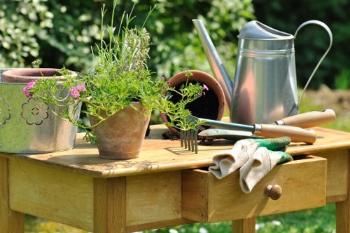 A scene of gardening tools and plant pots arranged on a wooden outdoor table in a lush garden setting. The table features a small drawer and is positioned amidst greenery with dense foliage in the background. On the table, there are two terracotta pots, one filled with a variety of green herbs and small flowering plants, and the other empty. Nearby, a metal watering can with a long spout and a wooden-handled garden fork are placed for outdoor gardening tasks. Also on the table are a pair of garden gloves, slightly worn, and a small trowel, ready for planting or soil preparation. The outdoor environment appears well-maintained, with natural light illuminating the scene, indicating a pleasant weather day suitable for garden work. The arrangement suggests ongoing gardening activity, relevant to landscaping and garden maintenance services provided by companies like Landscapers Forest Gate in the local area.