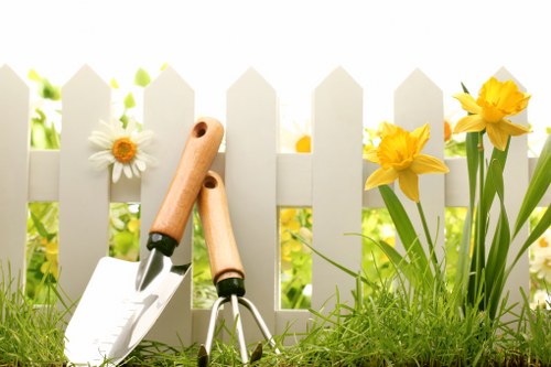 A close-up view of a well-maintained front garden with lush green grass and a variety of flowering plants, including yellow daffodils and white daisies, positioned along a white picket fence. The garden features a neat lawn area with dense, healthy grass and clear edges where soil and flower beds meet. Two gardening tools, a trowel and a hand rake, with wooden handles, are planted into the grass near the fence, suggesting recent gardening activity. In the background, the fence casts soft shadows, and daylight indicates clear weather conditions, creating a vibrant and inviting outdoor space typical of suburban gardens in the Forest Gate area. The scene emphasizes the importance of lawn care and planting work, relevant to professional gardening services offered by Landscapers Forest Gate, in maintaining and enhancing outdoor environments in UK towns and suburbs.