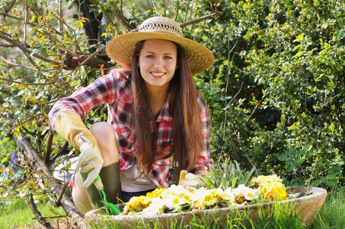 A young woman with long brown hair, wearing a wide-brimmed straw hat, a red and white checked shirt, and gardening gloves, is kneeling beside a flower bed in a garden. The flower bed contains blooming yellow and white flowers with dark centers, set within a curved, low brick edge. She is smiling while tending to the plants with a small garden tool in her right hand. The garden features lush green foliage, including leafy shrubs and trees in the background, with dappled sunlight filtering through the branches. The ground cover is a well-maintained grassy lawn, and a few fallen leaves and branches are visible nearby, indicating a tidy outdoor space. This scene captures a peaceful moment of outdoor gardening, highlighting natural textures, vibrant plant colours, and the garden’s structured layout, which may be part of a residential garden maintained by services such as those offered by Landscapers Forest Gate, serving the local area near the postcode E7 in London.