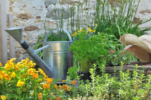 The image depicts a vibrant garden scene featuring a variety of flowering plants and greenery. In the foreground, there are bright yellow and orange flowers, possibly marigolds, alongside dense, green foliage. Behind these, taller plants with narrow leaves and small pink blossoms add textures and colour contrast. A metal watering can is positioned centrally among the plants, suggesting active garden maintenance. To the right, there is a wooden or wicker garden seat partially visible with a straw hat resting on it, indicating outdoor leisure or gardening activity. The background includes a weathered stone wall, adding a rustic charm to the outdoor space. The garden layout appears well-tended, with a mix of flowering borders and leafy shrubs, set in a temperate climate typical of the Forest Gate area, hinting at a tranquil, cultivated backyard. The lighting suggests a bright, sunny day, emphasizing the natural tones and lush greenery, aligning with gardening and landscape maintenance services offered by Landscapers Forest Gate.