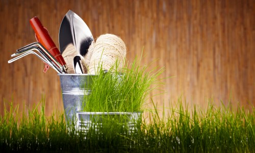 A close-up of a garden scene featuring a small patch of vibrant green grass in the foreground, with gardening tools and supplies placed on a metallic container. The tools include a trowel with a red handle, a pair of pruning shears, and a rake, alongside a ball of twine. Behind these items, a patch of slightly taller, healthy grass is visible, contrasting with the darker, textured wooden surface in the background, suggesting an outdoor setting suitable for garden maintenance and landscaping services in Forest Gate. The lighting appears natural, indicating a bright day, and the overall scene reflects a well-maintained outdoor space ready for gardening work.