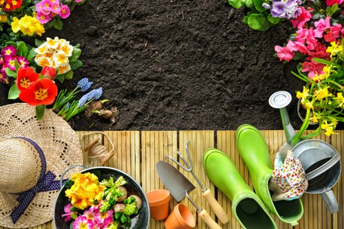 The image depicts a neatly arranged garden corner with a wooden decking area, bordered by a lush lawn with dense, green grass. In the upper section, there is exposed dark soil surrounded by vibrant flower beds featuring pink, purple, yellow, and red flowering plants, likely including primulas and pansies, creating a colourful border. On the decking, various gardening tools are neatly placed, such as green rubber boots, a small trowel, a hand fork, and a watering can, indicating recent gardening activity. Additionally, there are plant pots with blooming flowers, a straw sun hat with a navy ribbon, and a small orange watering can. In the background, the garden extends with more greenery and trees, suggesting a well-maintained outdoor space typical for residential gardens in Forest Gate, east London, offering a pleasant, inviting environment for outdoor gardening and relaxation. This scene highlights the garden’s layout with defined planting beds, practical tools, and vibrant plant diversity, matching the services offered by Landscapers Forest Gate in lawn and garden maintenance, landscaping, and decking repairs.