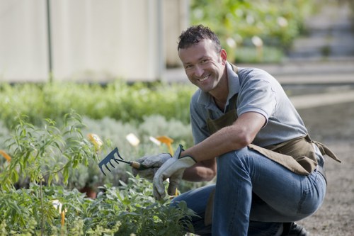 A man with dark, curly hair and a broad smile is working in a garden during daylight hours. He is kneeling on the soil, holding a gardening rake, and appears to be tending to or planting in a vegetable patch. The garden features a well-maintained lawn area with lush, green grass, bordered by a variety of plants and flowers. In the background, there is a wooden fence and a row of small trees or shrubs, suggesting a landscaped outdoor space, possibly part of a private garden or community area in Forest Gate. The scene is bright with natural sunlight, creating a cheerful and active outdoor environment. The man is dressed in casual gardening attire, including a grey shirt and jeans, with a brown apron or gardening vest, indicative of professional landscaping services offered by Landscapers Forest Gate. This setting highlights a typical, well-kept garden suitable for outdoor maintenance and landscaping improvements, reflecting local gardening and lawn care practices.